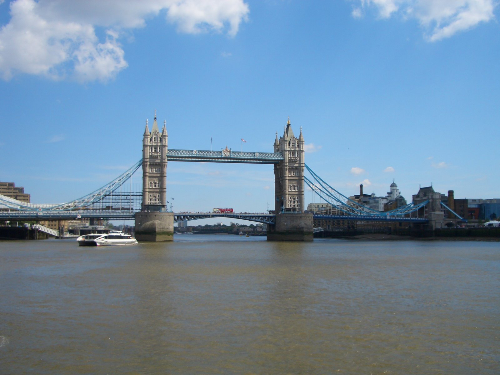 Tower Bridge spanning the River Thames on a bright day, with a white tour boat passing underneath and a blue sky with scattered clouds.
