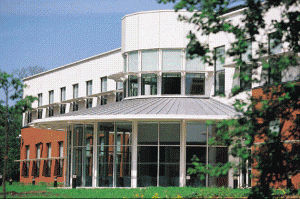 The Wellcome Trust Centre for Human Genetics, a modern building with a curved white upper storey, red-brick wings and a glass entrance under a conical roof, seen through trees.